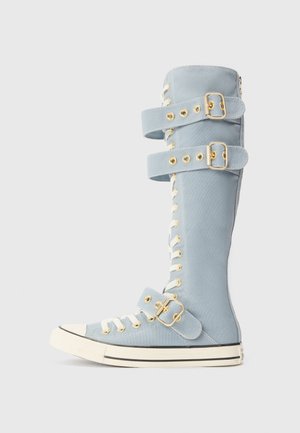 Světle modrá vysoká teniska s bílými tkaničkami, třemi nastavitelnými zlatými přezkami a bílou gumovou podrážkou s černým lemováním.