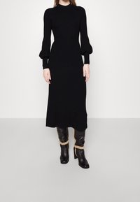 Schwarzes geripptes Strickkleid mit langen Puffärmeln und einer taillierten Silhouette, ergänzt durch knielange schwarze Stiefel mit tanfarbigen Akzenten.