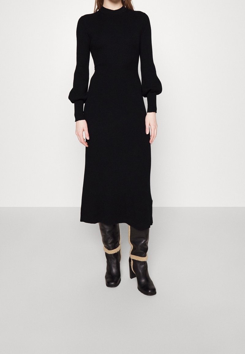 Schwarzes geripptes Strickkleid mit langen Puffärmeln und einer taillierten Silhouette, ergänzt durch knielange schwarze Stiefel mit tanfarbigen Akzenten.