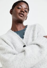 Cardigan grigio in maglia con una texture morbida, caratterizzato da un profondo scollo a V, polsini a costine e una chiusura con un singolo bottone. Indossato sopra una maglietta teal.
