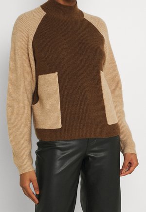 Gebreide trui met hoge kraag, in bruin en beige kleuren, geribbelde textuur en twee opgestikte zakken; gedragen met zwart leren broeken.