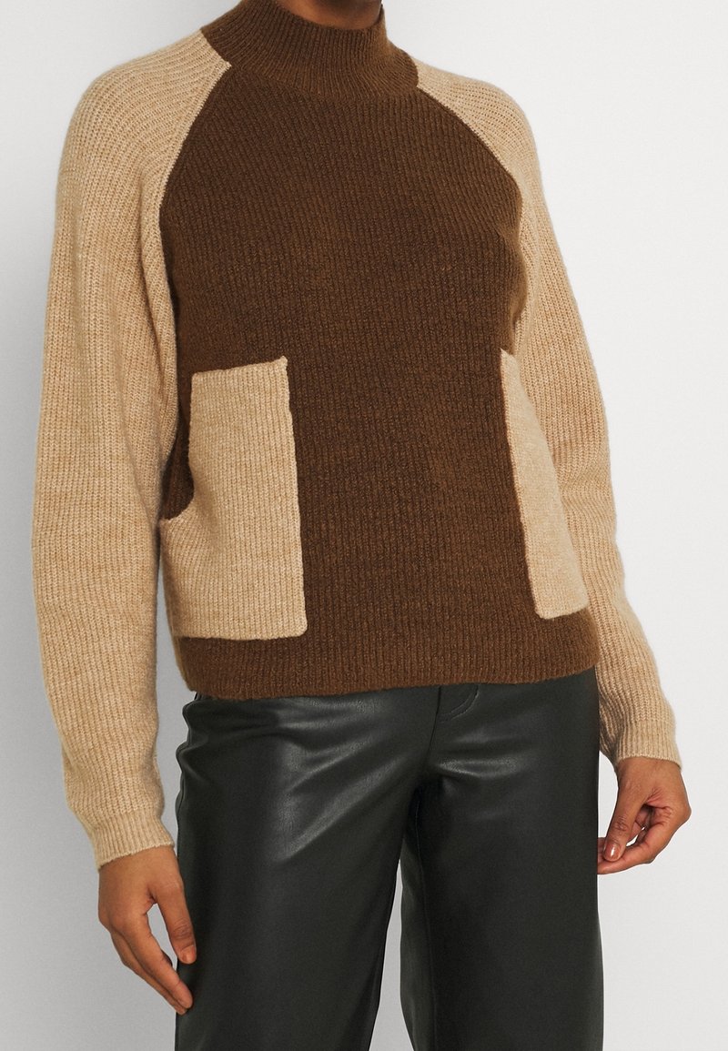 Strickpullover mit hohem Kragen, in Braun- und Beigetönen, mit gerippter Textur und zwei aufgesetzten Taschen; kombiniert mit schwarzen Lederhosen.