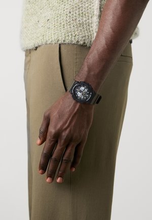 Montre numérique noire au poignet avec un bracelet en caoutchouc texturé, dotée d'un cadran rond affichant plusieurs cadrans, le tout sur un vêtement vert discret.