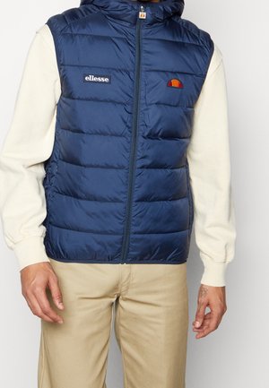 Mand iført navy blå puffer vest med ellesse-logo over creme sweatshirt og beige bukser, stående imod en ensfarvet baggrund.