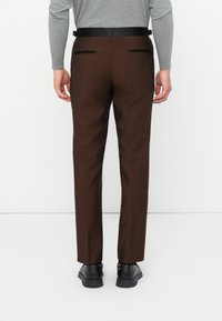 Bărbat îmbrăcat cu pantaloni eleganți maro cu buzunare pe spate, un top gri cu mânecă lungă, pantofi negri și o curea neagră, stând pe un fundal alb.