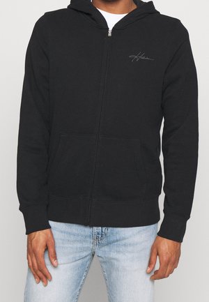 Homme portant un sweat à capuche noir zippé avec poches avant et petit logo sur la poitrine, associé à un jean bleu clair.