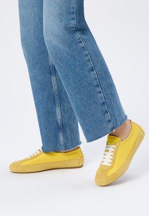 Personne portant des baskets jaunes à lacets avec des accents en suède et un jean bleu raccourci, debout sur un fond blanc uni.