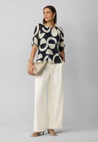 Top plissé navy et crème présentant de grands motifs circulaires, associé à un pantalon large blanc et un sac beige avec des accents en chaîne.