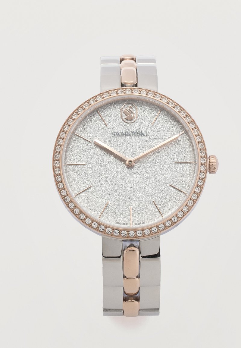 Στρογγυλό ρολόι Swarovski με καντράν ασημί με γκλίτερ, δείκτες σε ροζ χρυσό, στεφάνη διακοσμημένη με κρυστάλλους και δίχρωμο μεταλλικό λουράκι που δείχνει 10:10.