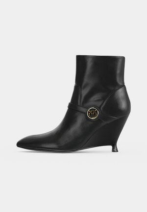 Bottine noire en cuir avec bout pointu, talon compensé et boucle circulaire décorative dorée et argentée sur la bride latérale.