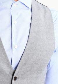 Gilet grigio realizzato in materiale testurizzato sopra una camicia azzurra chiara con piccole pois blu, caratterizzato da un bottone marrone e colletto bianco.