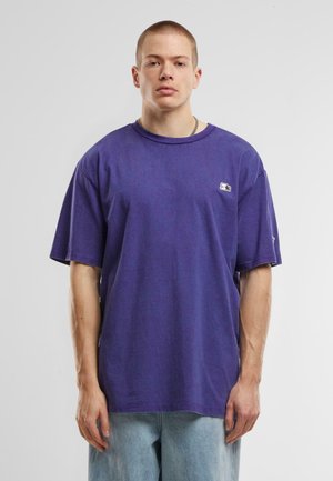 Maglietta di cotone viola con una vestibilità rilassata, maniche corte e un piccolo logo ricamato sul petto. Indossata con jeans azzurri larghi.