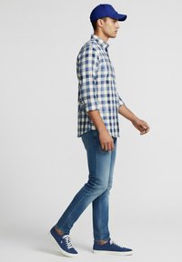 Hombre caminando, con gorra de béisbol azul, camisa a cuadros azul y blanca, jeans ajustados azules y zapatillas azules con cordones blancos.