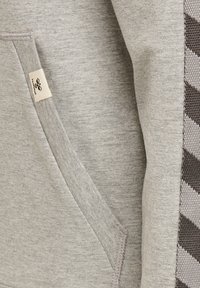 Sudadera gris claro con un bolsillo frontal, tela texturizada y un panel lateral con un patrón de zigzag en marrón oscuro.