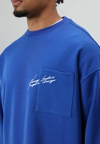 Blauwe trui met een ronde hals en een zakje op de borst. Heeft witte geborduurde tekst "Young Leaders Inspire Change". Glad oppervlak.