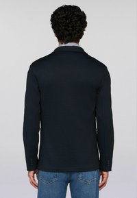 Muž s kudrnatými vlasy, oblečený v černém sakou přes pruhovanou košili a modré džíny, stojící zády k jednoduchému šedému pozadí.