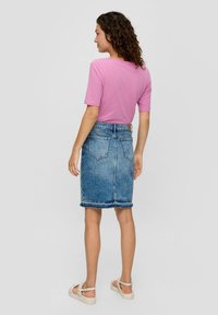 Vrouw met krullend haar, gekleed in een roze top met korte mouwen, een blauwe denim rok tot de knie en beige sandalen, staand en met de rug naar de kijker gekeerd.