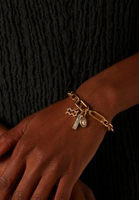 Gouden kettingarmband met gevarieerde schakel, met bedels: een rechthoekige witte steen en een ovale heldere steen versierd met kleine kristallen.