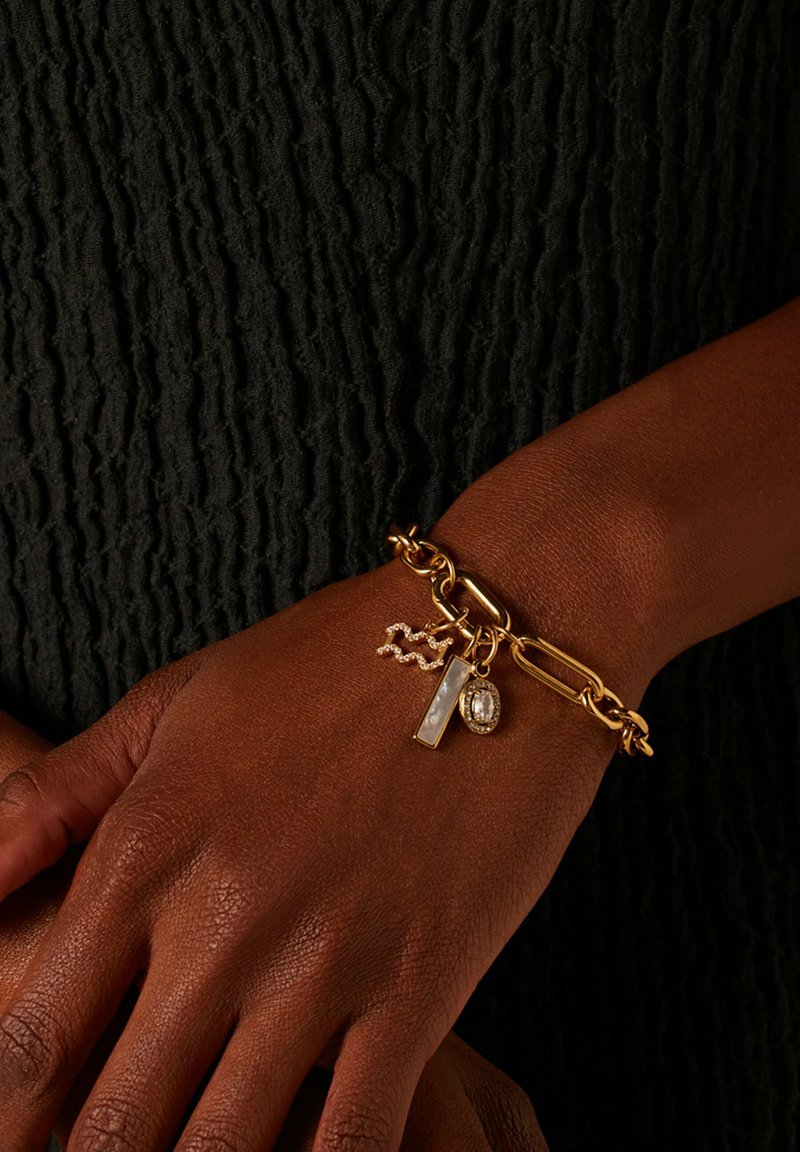 Gouden kettingarmband met gevarieerde schakel, met bedels: een rechthoekige witte steen en een ovale heldere steen versierd met kleine kristallen.
