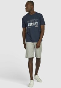 Marineblauwe t-shirt met de tekst "CITY ESCAPE" graphic, gecombineerd met lichtgrijze shorts en witte sneakers, staand tegen een effen achtergrond.