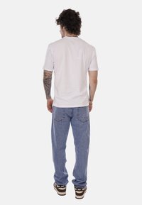 Uomo con tatuaggi che indossa una maglietta bianca a maniche corte semplice, jeans blu e sneakers Nike marroni, in piedi di spalle su uno sfondo bianco.