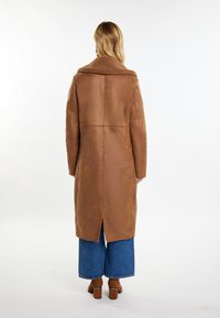Brun faux læderfrakke med en tekstureret, plysset krave; lang, lige design; rygslids; båret over brede denimbukser.