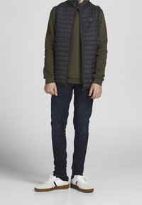 Child wearing dark puffer vest over olive sweatshirt, dark skinny jeans, and white sneakers standing on a plain gray background.