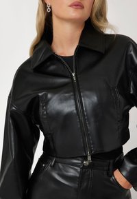 Juodas odinis trumpas švarkas su užtrauktuku, dideliu apykakliu ir lygiu paviršiumi. Priekyje yra kišenės. Suderinta su atitinkamomis kelnėmis.