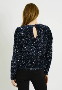 Top azul marino de manga larga con lentejuelas y tejido texturizado, con diseño de espalda con abertura en forma de gota y escote redondo. Combinado con pantalones negros ajustados.