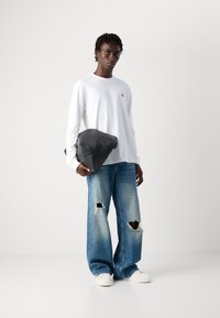 Hvit skjorte med lange ermer og en liten logo, kombinert med løse blå jeans med slitte knær og hvite sneakers; holder en svart lærveske.