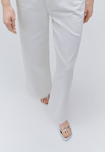 Pantalon blanc à jambes larges porté avec des talons argentés à brides et orteils ouverts, montrant les jambes jusqu'aux mollets et les mains détendues sur les côtés.
