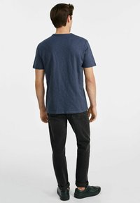 Granatowa koszulka z okrągłym dekoltem, krótkimi rękawami i luźnym krojem, zestawiona z czarnymi dżinsami i zielonymi trampkami.