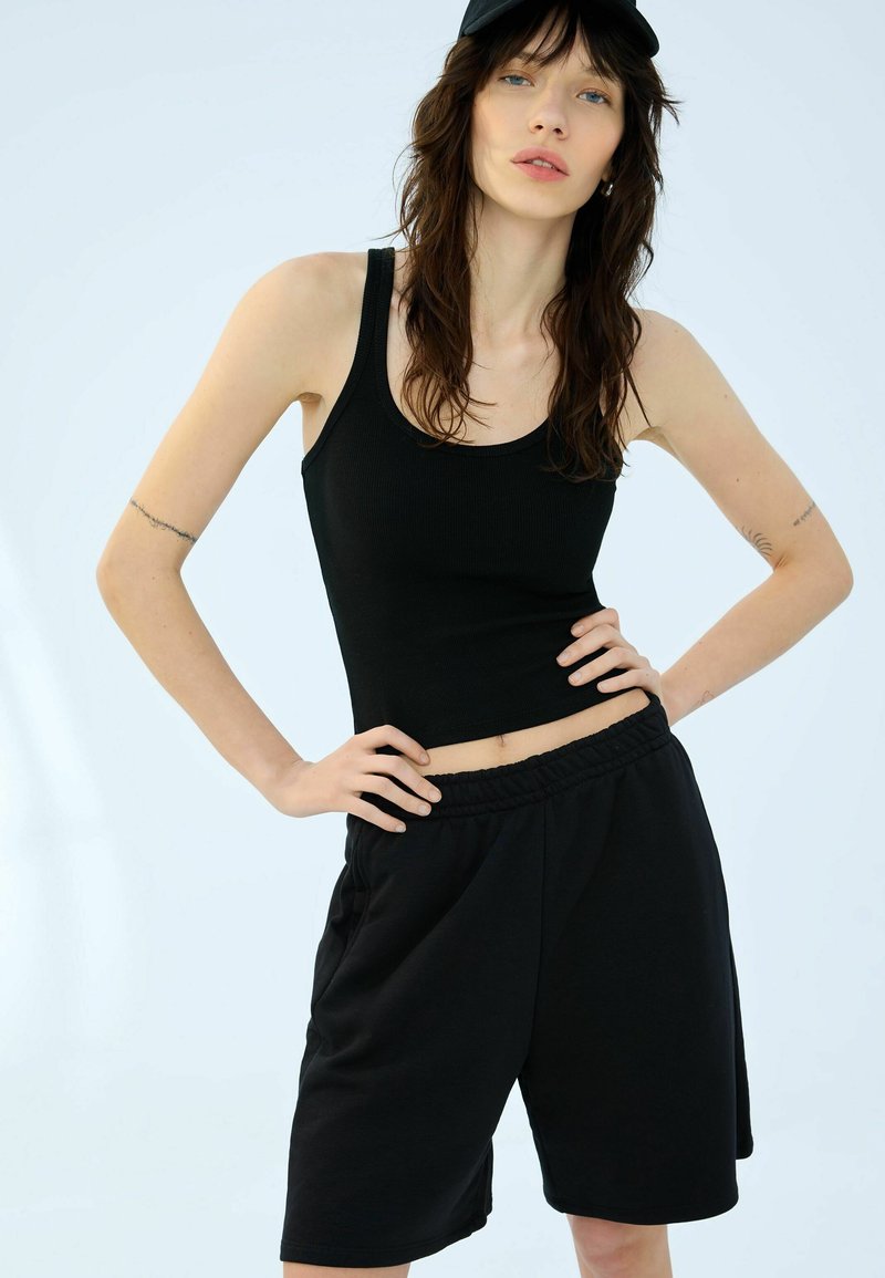 Čierny rebrovaný tielko a čierne šortky s elastickým pásom. Tkanina je ľahká s hladkou textúrou. Model pózuje s rukami na bokoch.