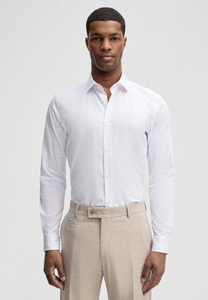 Mężczyzna stojący przodem, ubrany w białą koszulę i beżowe, teksturowane spodnie, na jednolitym tle.