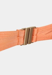Artimiausias auksinės spalvos metalo sagties užsegimas, jungiantis du oranžinės spalvos audinio juosteles baltoje fone.
