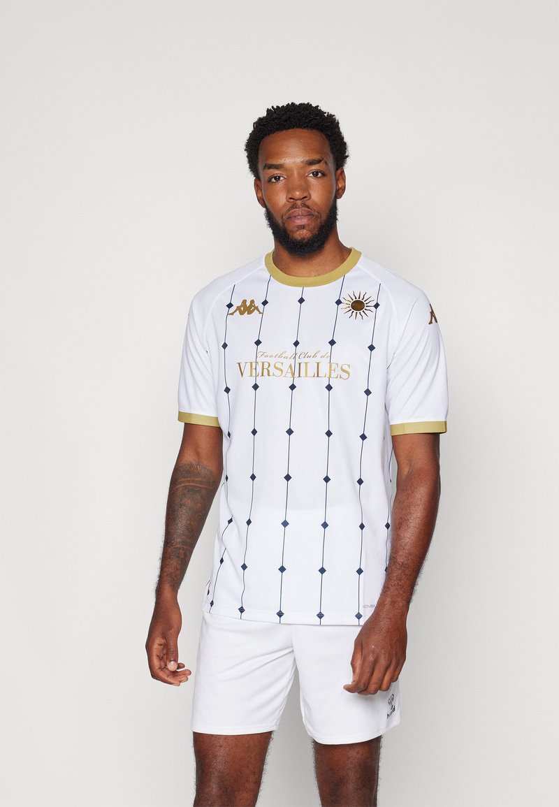 Vit fotbollströja med gyllene detaljer och marinblå diamantmönster. Har en rund hals och broderad logotyp. Kombineras med vita shorts.