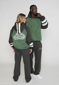 Hanorace cu glugă, în verde și negru, cu dungi albe; fermoar frontal, buzunare laterale și text cu logo distinctiv pe spate. Asortate cu pantaloni închisi la culoare.