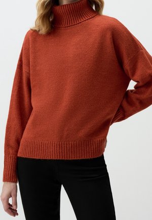 Femme portant un pull à col roulé tricoté couleur rouille avec des poignets et un ourlet côtelés, associé à un pantalon noir sur un fond uni.