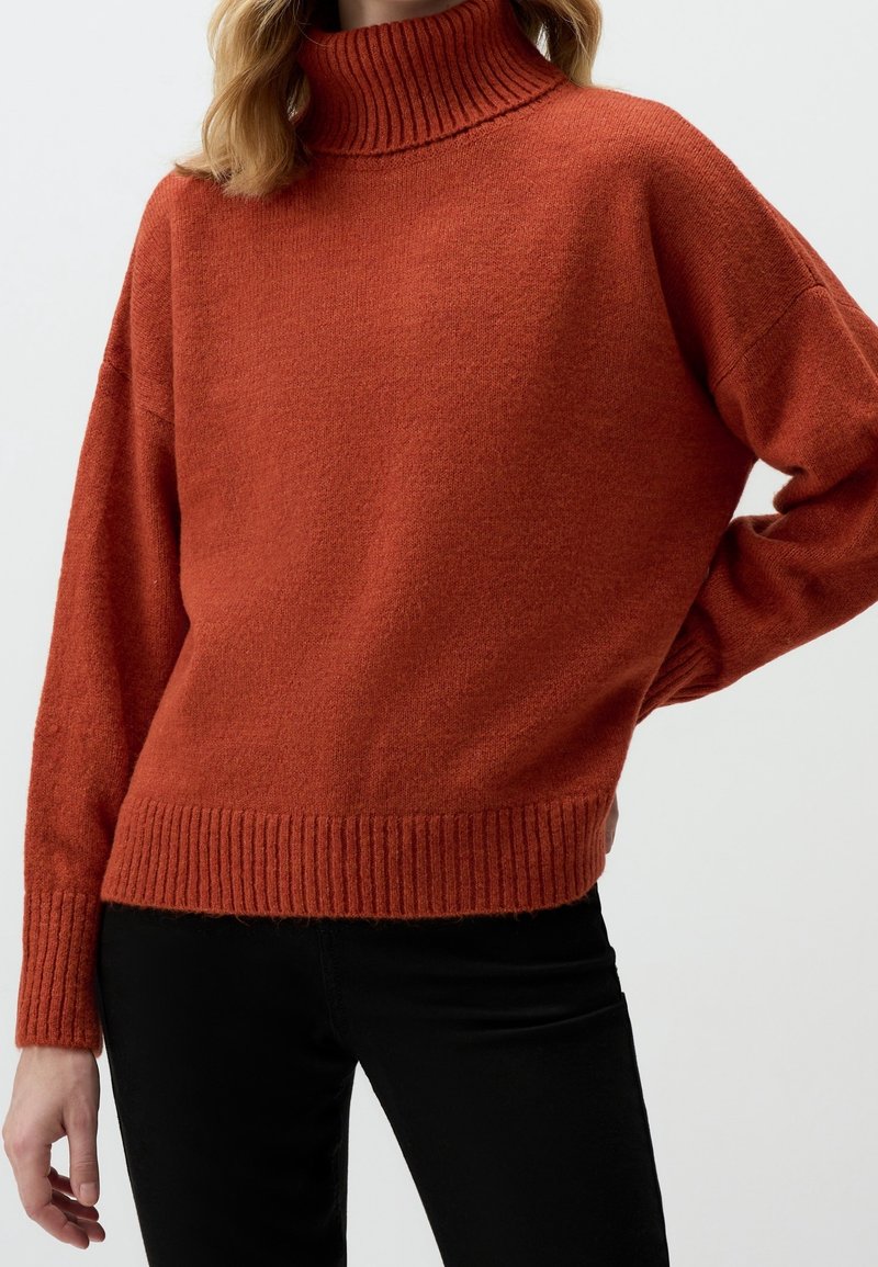 Femme portant un pull à col roulé tricoté couleur rouille avec des poignets et un ourlet côtelés, associé à un pantalon noir sur un fond uni.