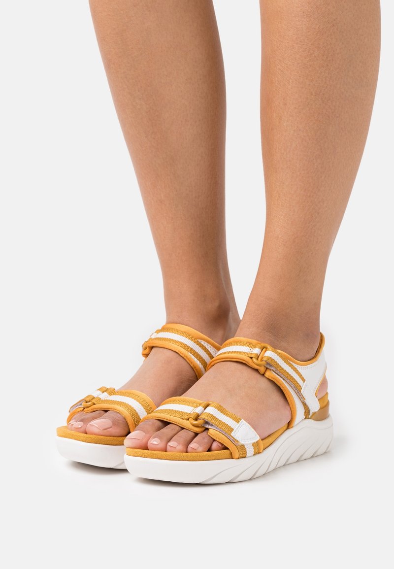 Pieds portant des sandales sportives blanches et jaune moutarde avec des sangles rayées réglables et des semelles blanches épaisses, debout sur une surface claire.