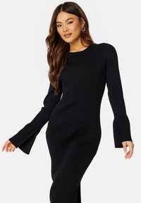 Abito nero a coste in maglia con maniche lunghe, polsini svasati e uno spacco laterale. Presenta un colletto rotondo e una silhouette aderente.