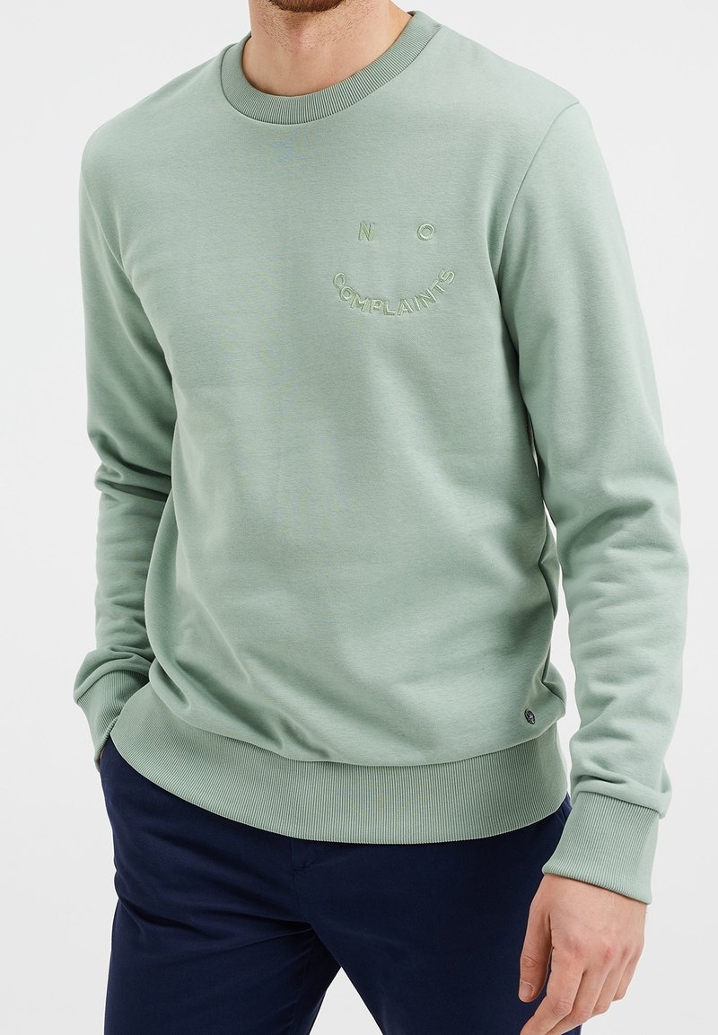 Muž oblečený v svetle zelené mikině s nápisem "ŽÁDNÉ STÍŽNOSTI" vyšítým na hrudi, v tmavě modrých kalhotách, s rukama u těla.