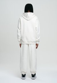 Sweat-shirt blanc à capuche avec poignets élastiques, associé à un pantalon de jogging blanc assorti, vu de dos. Les baskets présentent des accents noirs et blancs.