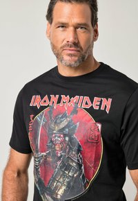 Maglietta nera in cotone con grafica colorata di uno scheletro samurai, evidenziata da accenti rossi, dettagli intricati e testo audace.