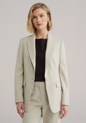 Lysebeige blazer med struktureret design, enkelt knaplukning og to forlommer. Båret over en mørk blonder-top. Glat tekstur.