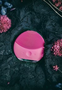 Appareil de nettoyage facial en silicone rose avec surface texturée, forme arrondie et boutons de contrôle, posé sur de la dentelle noire et des fleurs.
