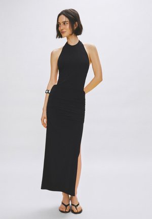 Femme aux cheveux courts et foncés, portant une robe longue noire sans manches avec un décolleté halter et une fente latérale, ainsi que des sandales noires, debout avec une main dans le dos.