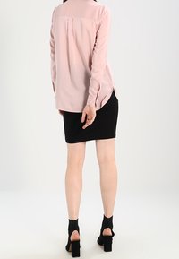 Langärmlige, blassrosa Bluse mit einer Rückenfalte, kombiniert mit einem enganliegenden schwarzen Rock. Das Modell trägt schwarze Pumps mit Knöchelriemen.