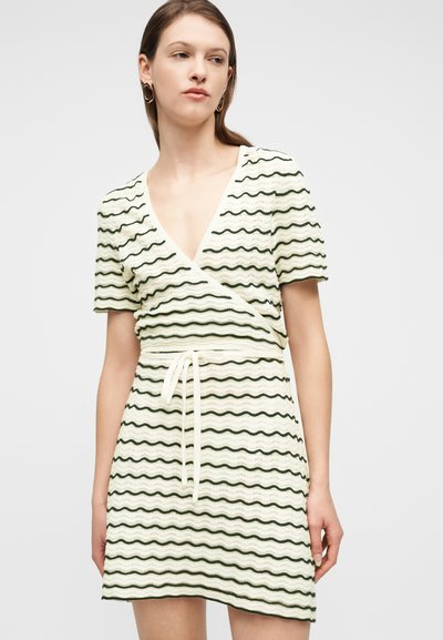 Γυναίκα που φοράει φόρεμα σε εκρού χρώμα με κοντά μανίκια, V λαιμόκοψη, πράσινες κυματιστές ρίγες και δεμένη ζώνη στη μέση, στέκεται μπροστά σε απλό λευκό φόντο.