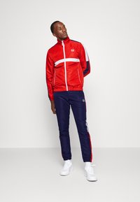 Chaqueta deportiva roja con franja blanca y detalles en azul marino, combinada con pantalones azul marino. Ambos hechos de un material suave y ligero con puños elásticos.